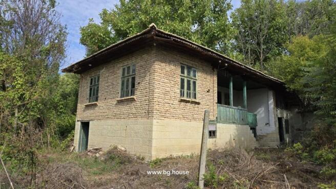 View of a traditional Bulgarian house showing stone lower walls and wooden upper walls with green window frames, surrounded by trees and garden vegetation.
