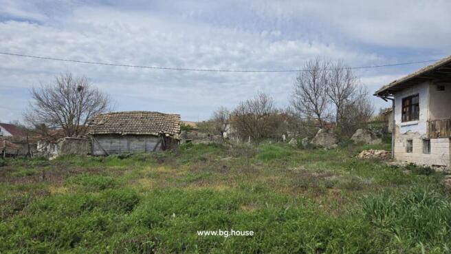 View of the garden area with wild grass and purple flowers adjacent to a traditional Bulgarian house with stone basement and tiled roof.