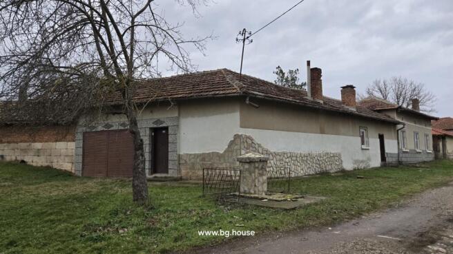 Corner view of a Bulgarian house featuring stone and plaster exterior walls, a metal gate, and a leafless tree in front.