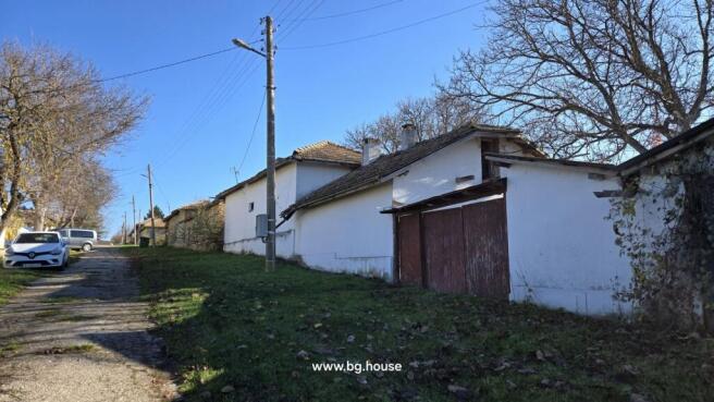 View along the village street showing the white cottage, adjacent outbuildings, parked cars, and a utility pole.