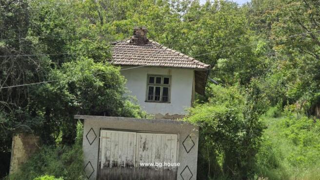 Close-up view of the old Bulgarian house showing the tiled roof, garage door, and surrounding green bushes.