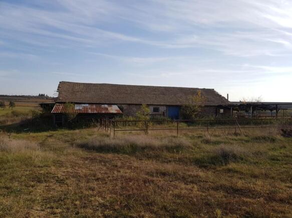 View of the long farm building with a tiled roof and metal fencing in front, surrounded by dry grass and open countryside.