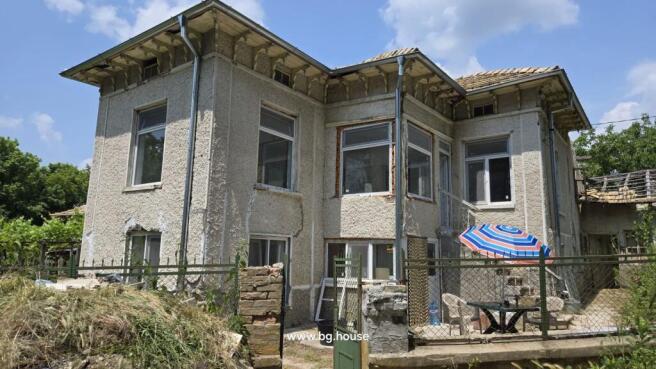 Side perspective of the Bulgarian house highlighting the textured walls, windows, and fenced yard with table and chairs under a striped umbrella