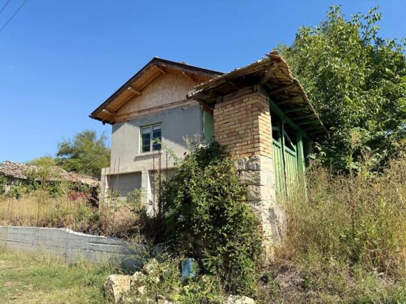 Side perspective of the old house with surrounding overgrown plants and a small brick outbuilding with tiled roof.