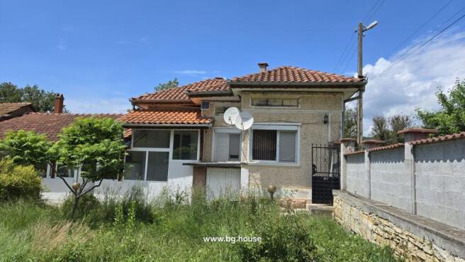 Exterior view of a Bulgarian house with terracotta tiled roof, satellite dishes, and a garden with green trees and grass.