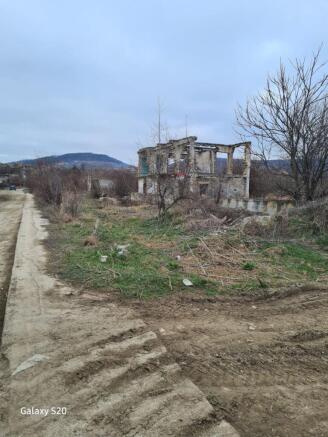 A ruined two-story concrete building frame stands on a grassy plot with bare trees and a dirt road beside it under a cloudy sky.