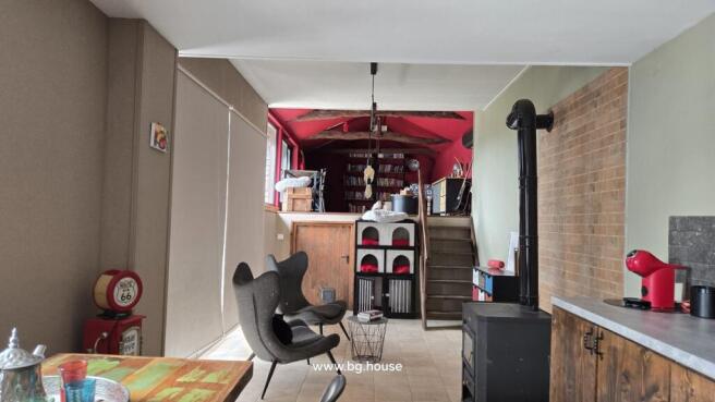 Interior living space showing two modern chairs, a wood stove, and exposed red ceiling beams with bookshelves in the background.