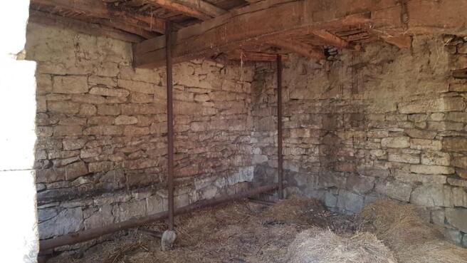 Inside view of a stone outbuilding showing wooden ceiling beams and metal poles supporting the structure, with straw covering the floor.