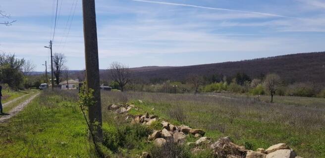 A dirt road runs alongside a grassy field bordered by a stone wall and utility pole, with trees and hills in the distance.