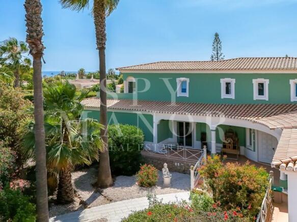 Exterior view of a green Algarvian luxury villa in Carvoeiro, showcasing traditional architecture, a paved garden path, palm trees, and spacious covered terraces.