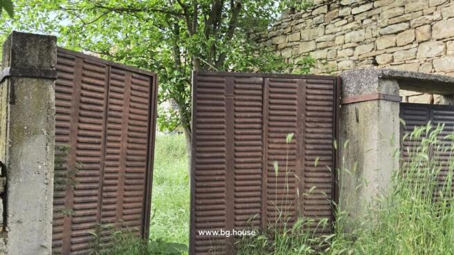 Close-up of a rusty metal gate next to a stone wall with green plants growing around it.