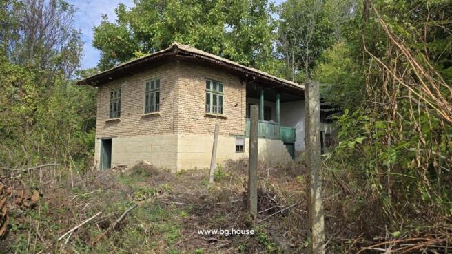 Side perspective of a traditional Bulgarian house featuring a stone cellar, wooden balcony with green railings, and garden area with trees and shrubs.