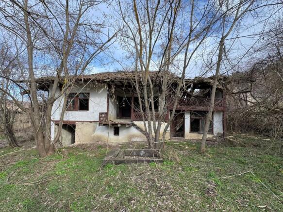 Exterior view of an old house in Balkanski village showing white walls, wooden balcony, and surrounding bare trees.