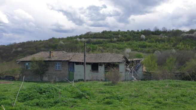 Side perspective of an old house showing a partially collapsed roof and green field with hills in the background.