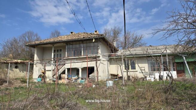 View of a two-storey Bulgarian house with a tiled roof, external stairs, and a garden area with dry grass and leafless trees