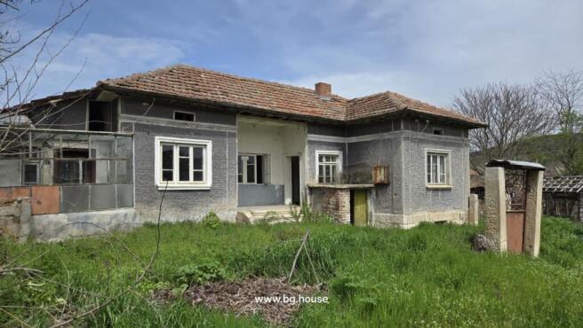 Front view of the Bulgarian rural house showing grey walls, tiled roof, and surrounding garden area