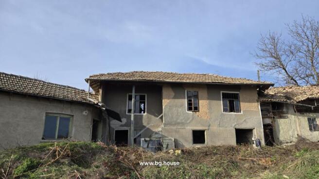 Exterior view of an old Bulgarian house with tiled roof and several windows, showing the rustic facade and surrounding dry vegetation.