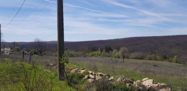View of a grassy rural field bordered by a stone wall and a utility pole, with hills in the background under a partly cloudy sky.