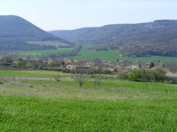 Green fields with scattered trees and a village nestled at the base of forested hills