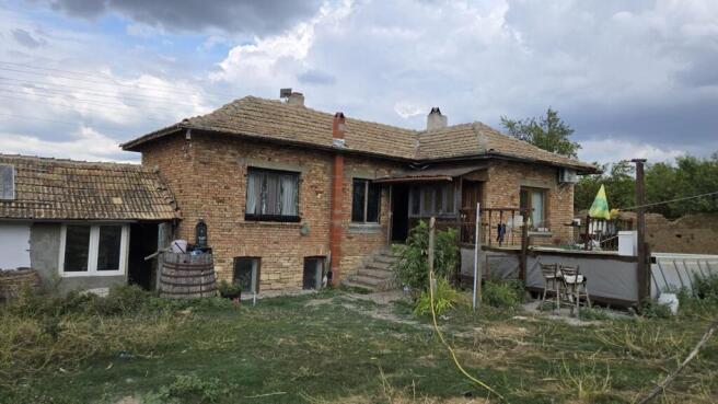 Exterior view of a brick house with a tiled roof, wooden porch, and garden area with grass and plants.