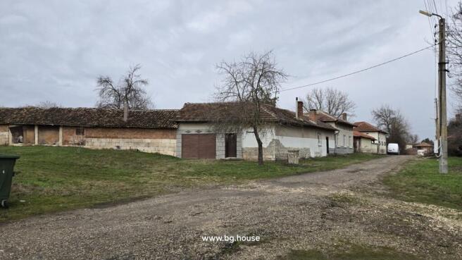 View of a corner Bulgarian house with tiled roof, stone base, and adjacent buildings at the end of a village street.