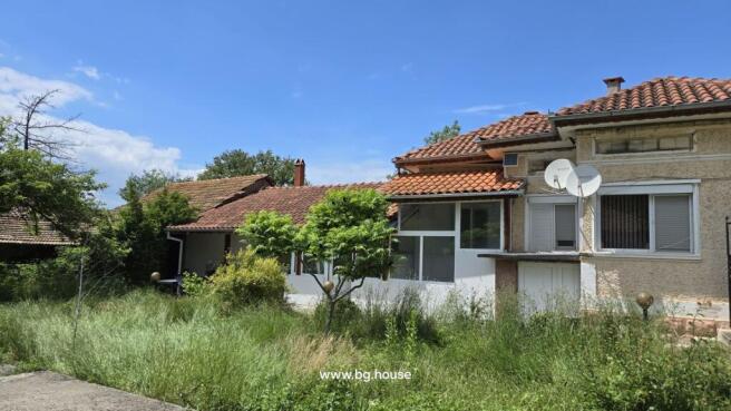 Side perspective of the Bulgarian house showing tiled roof, enclosed garden with tall grass and small trees.