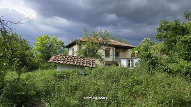 View of the yellow two-storey house with a balcony, surrounded by dense green vegetation and a cloudy sky.