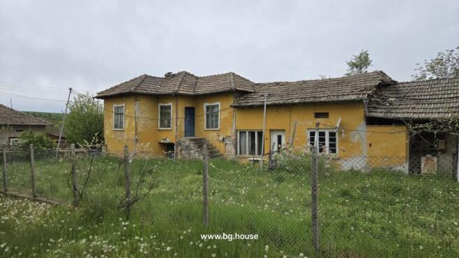 Front view of a yellow rural house with a tiled roof and stone steps leading to the entrance, enclosed by a wire fence with green grass and dandelions