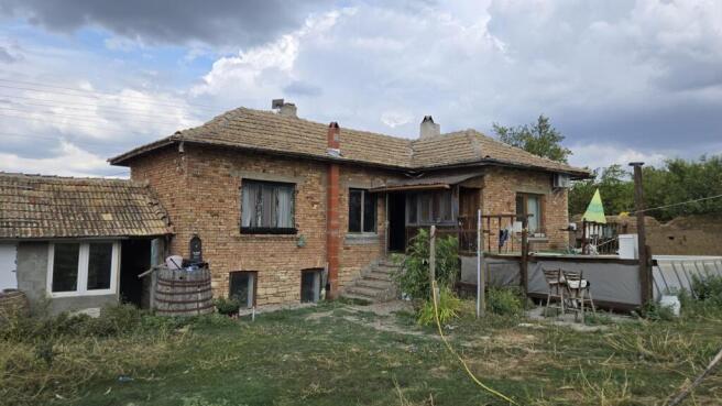 Exterior view of a brick house with a tiled roof, wooden porch, and garden area with grass and plants.