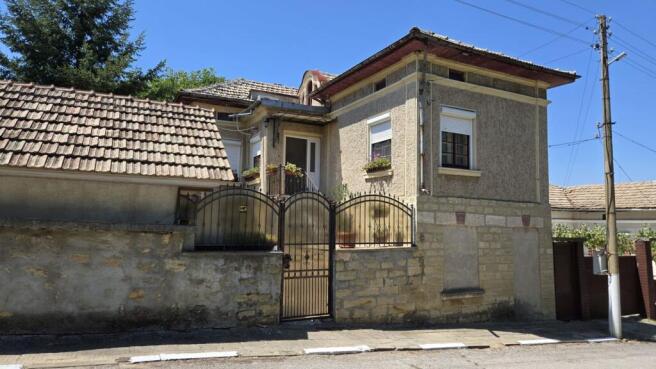 Front view of a traditional Bulgarian house with a tiled roof, stone wall, and metal gate.