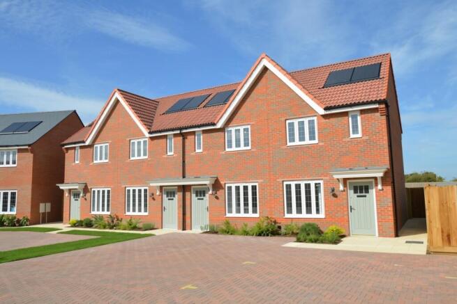 Modern red-brick houses with solar panels stand behind a paved driveway and neatly trimmed green law