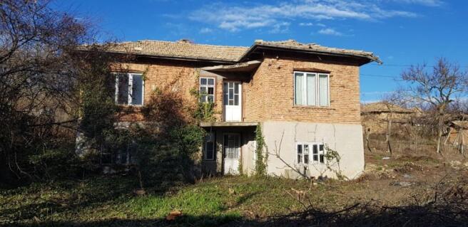 Front view of a two-storey rural Bulgarian house made of brick and stone with a tiled roof and surrounding trees.