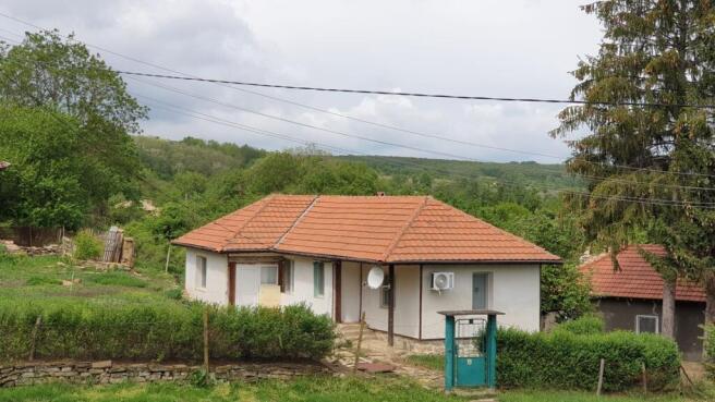 A white single-storey house with a terracotta tiled roof set in a green rural environment with trees and bushes.