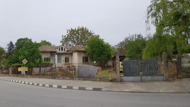 View of a single-storey house with a tiled roof surrounded by a wire fence and trees on a street corner