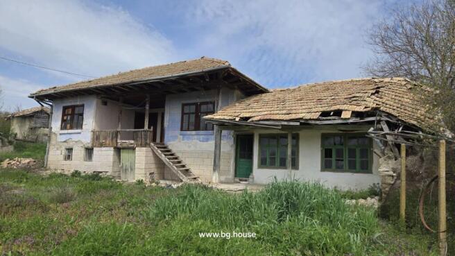 Exterior view of a traditional Bulgarian house showing stone basement, wooden balcony, tiled roof, and green doors and windows.