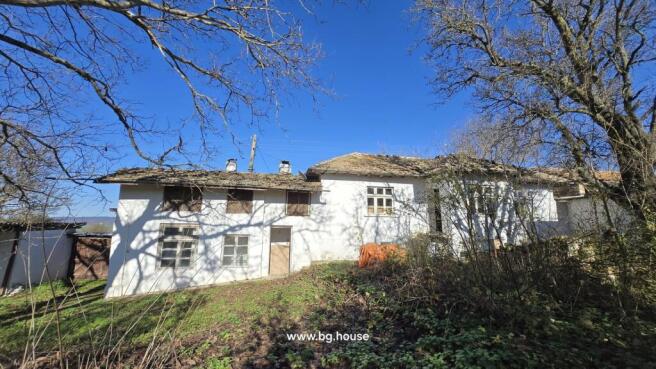 View of the white two-storey Bulgarian cottage at the end of the village with leafless trees casting shadows on the facade.