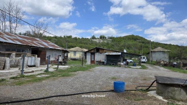 View of multiple farm buildings and a yard area with gravel and grass, surrounded by trees and hills.