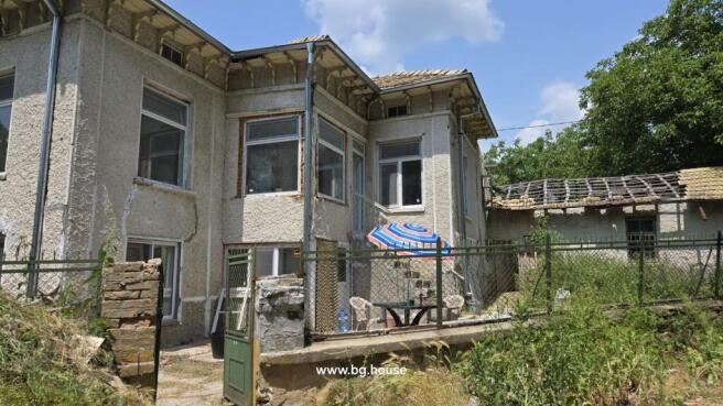 View of the Bulgarian house from a different side angle showing the textured walls, multiple windows, and fenced yard with outdoor seating