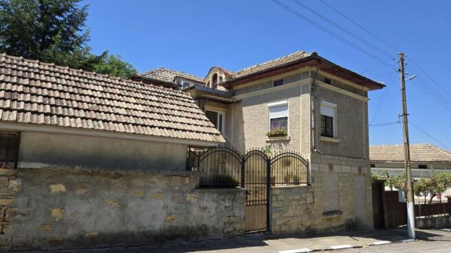 Side perspective of the Bulgarian house highlighting the tiled roof and stone wall with a metal gate.