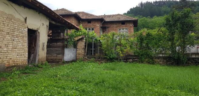 View of a brick house with tiled roof and green garden area enclosed by stone and wire fencing, with forested hills in the background.