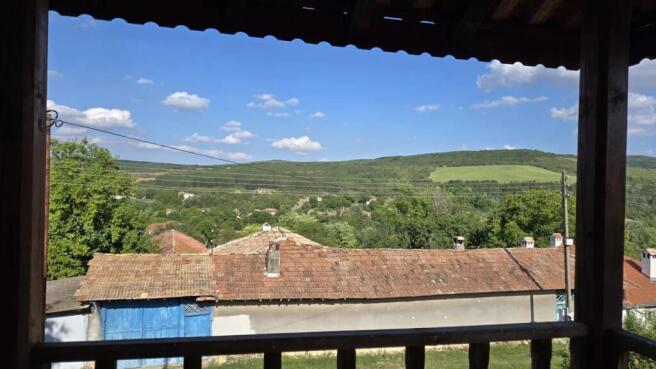 View from a covered wooden balcony overlooking village rooftops and green hills under a partly cloudy blue sky.