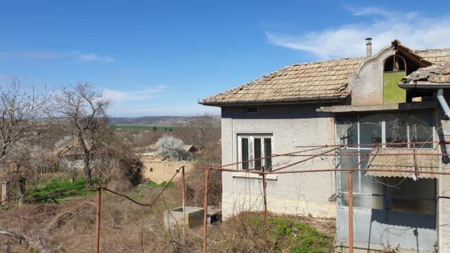 View of a village house with a tiled roof and enclosed porch, overlooking a garden area with both dry and green vegetation patches.
