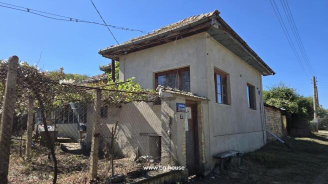 Exterior of a two-storey village house with plastered walls and wooden windows, featuring a fenced yard covered with grapevines