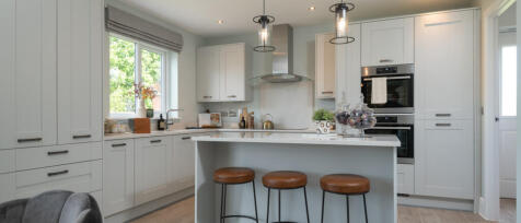 Modern white kitchen with island, pendant lighting, and large window showing green trees outside.