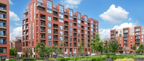 Modern red brick residential buildings with balconies, surrounded by greenery and a bright blue sky