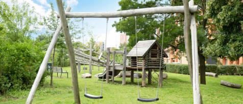 A wooden children&#39;s play area with swings, a slide, and a small house in a green park with trees and