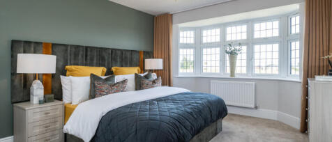 A cosy bedroom with a large bay window, a double bed with colourful cushions, and modern bedside lam