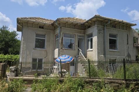 Front view of the two-storey Bulgarian house showing textured walls, multiple windows, and a small outdoor seating area with a striped umbrella