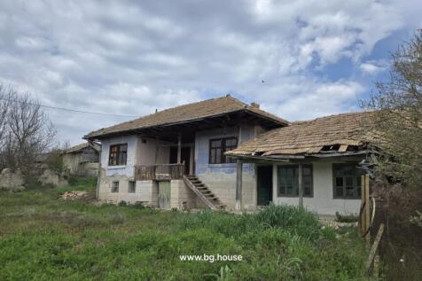 Exterior view of a traditional Bulgarian house showing stone basement, wooden balcony, and tiled roof surrounded by grass.