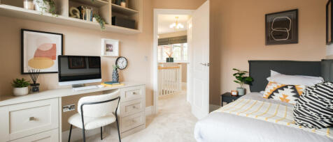 A cosy bedroom with a white desk, computer, artistic wall frames, a bed with patterned cushions, and